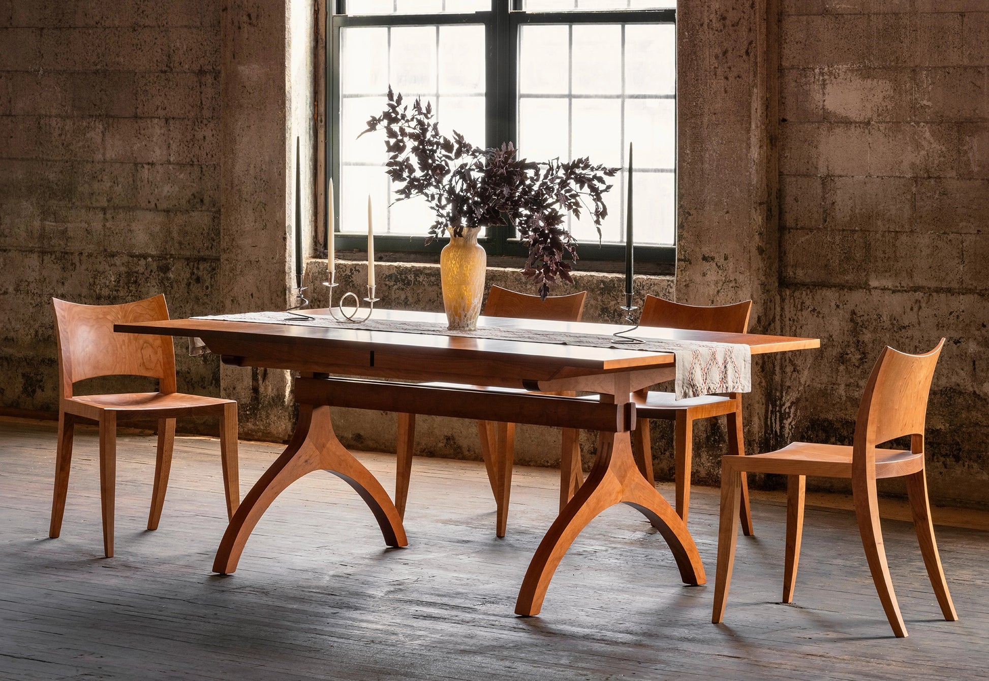 Wooden dining table with chairs in a room with large windows and concrete walls.