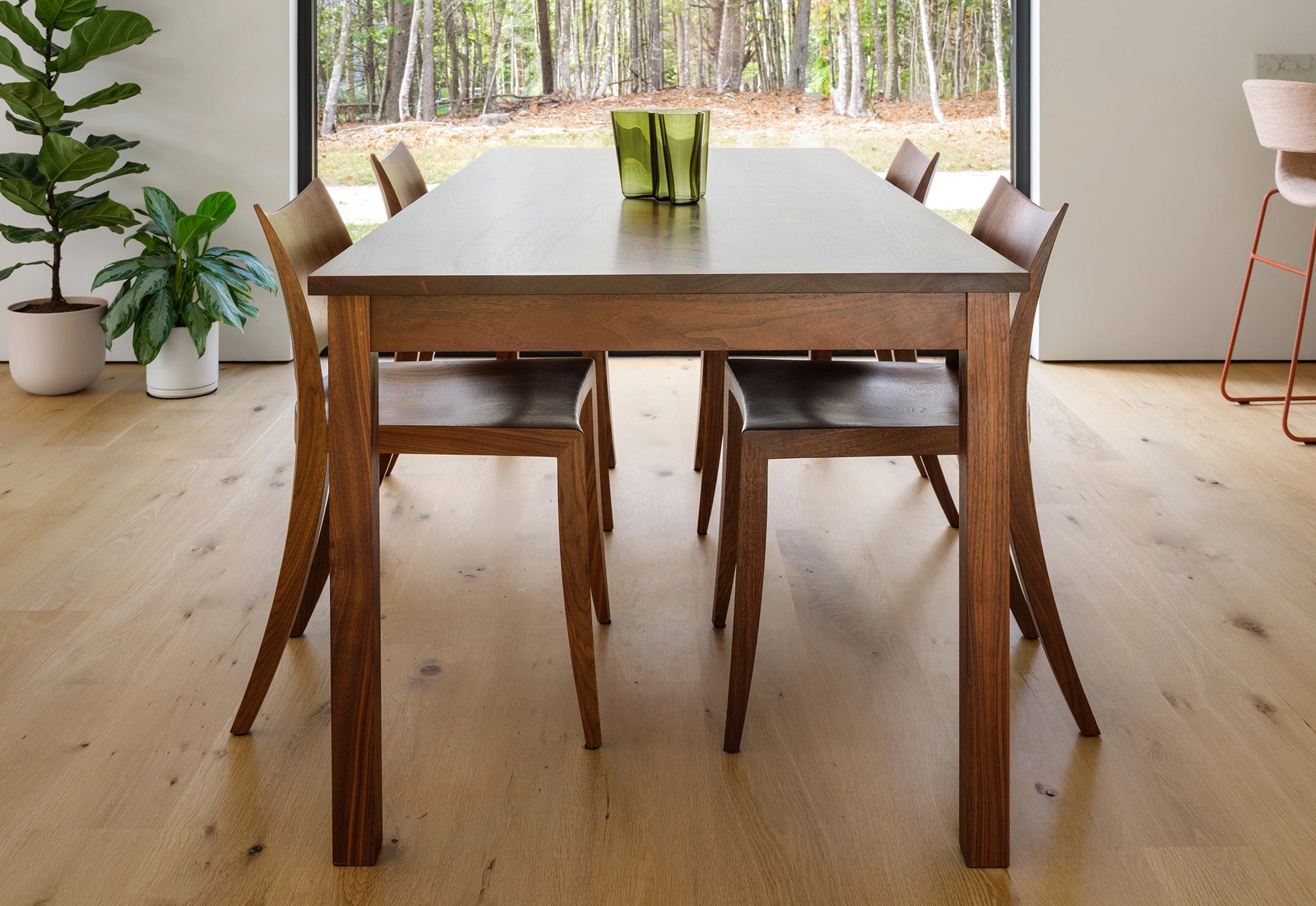 Wooden dining table with chairs in a room with large windows showing a forest view.