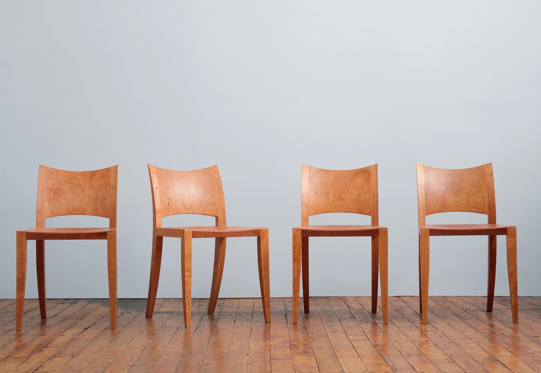 Four wooden chairs lined up against a plain wall on a wooden floor.