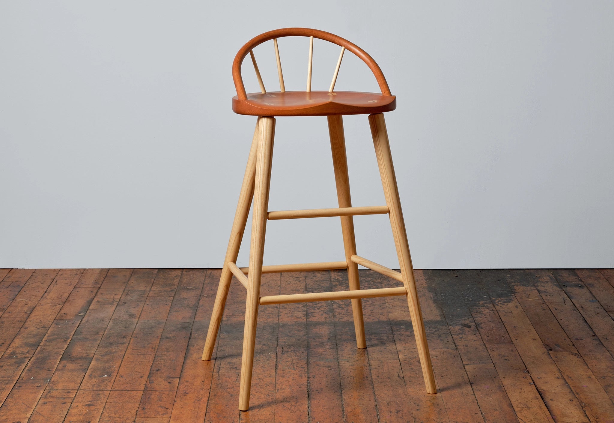 Wooden bar stool with a curved backrest on a wooden floor against a gray wall.