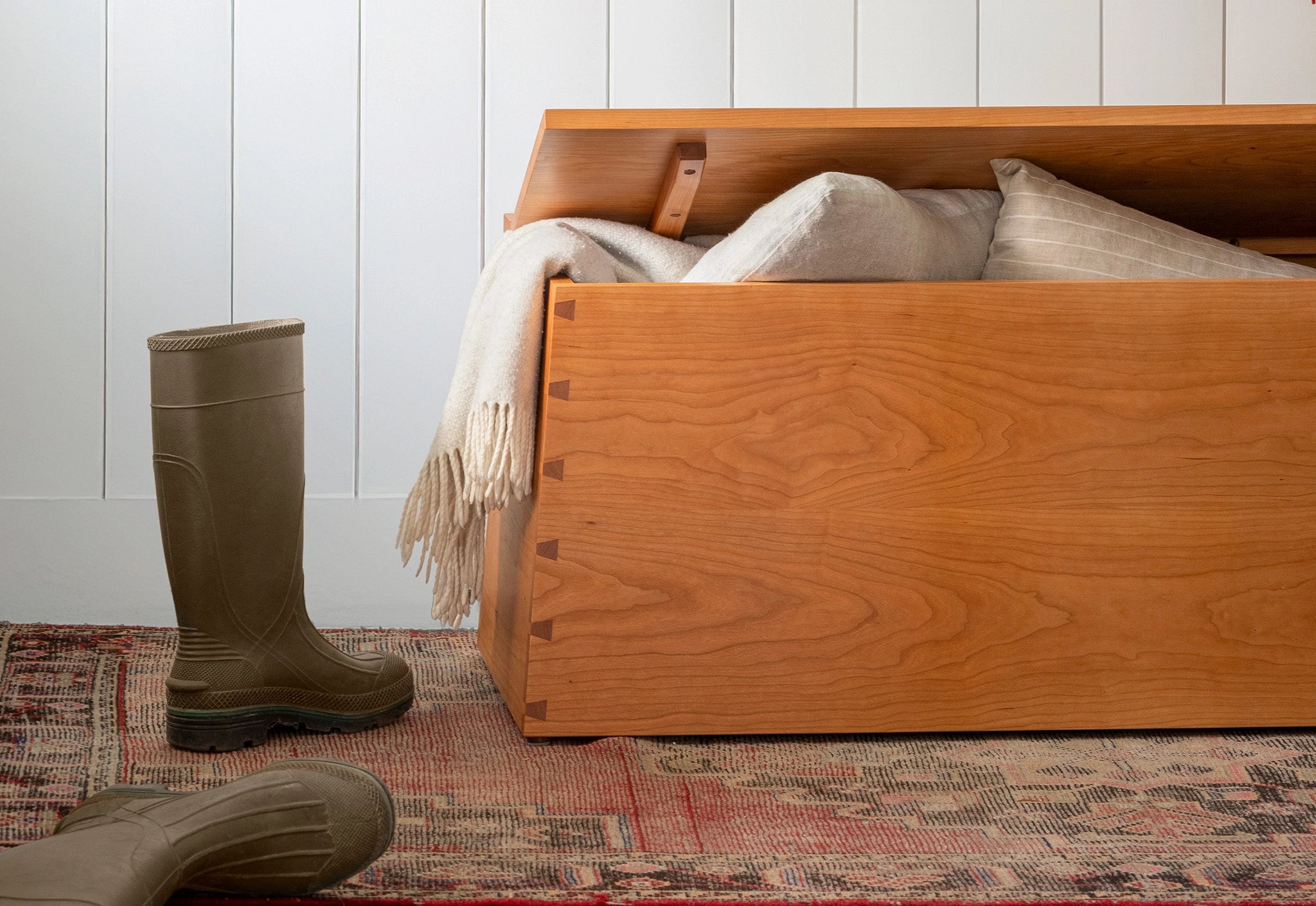 Wooden storage chest with a blanket and boots on a patterned rug against a white paneled wall.