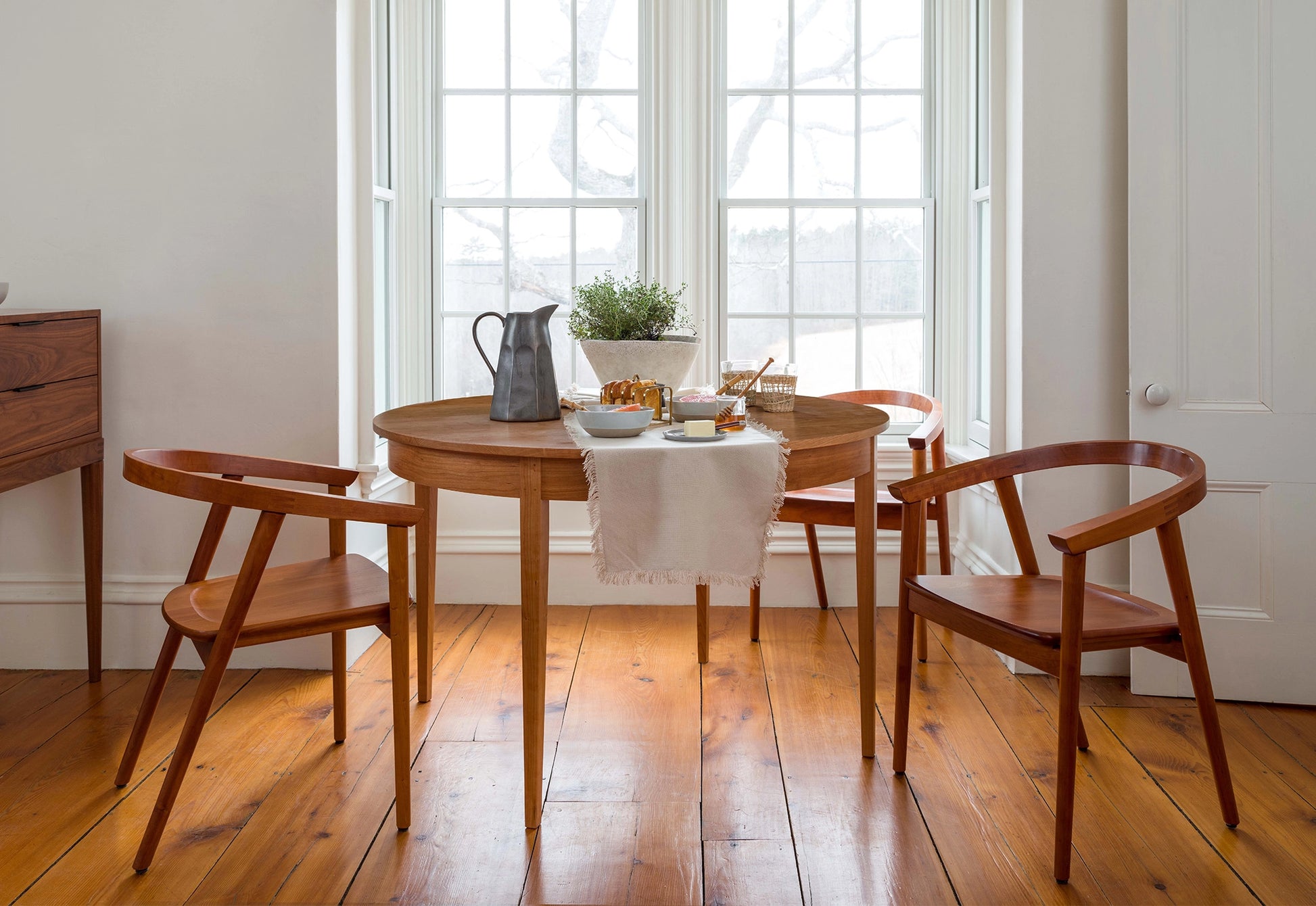 Wooden dining table with chairs in a bright room with large windows.