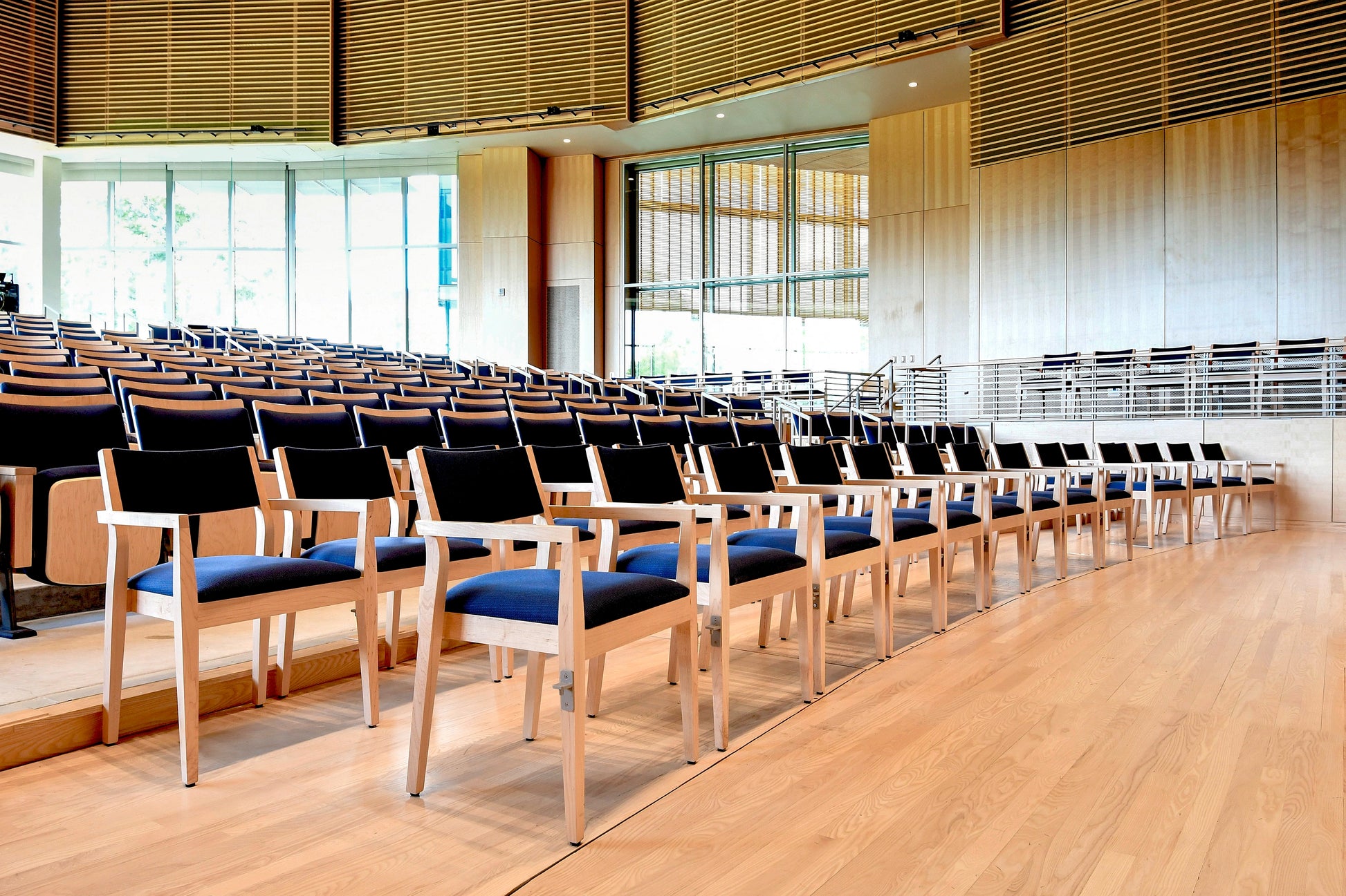 Row of chairs with blue cushions in a large, well-lit room with wooden flooring and large windows.