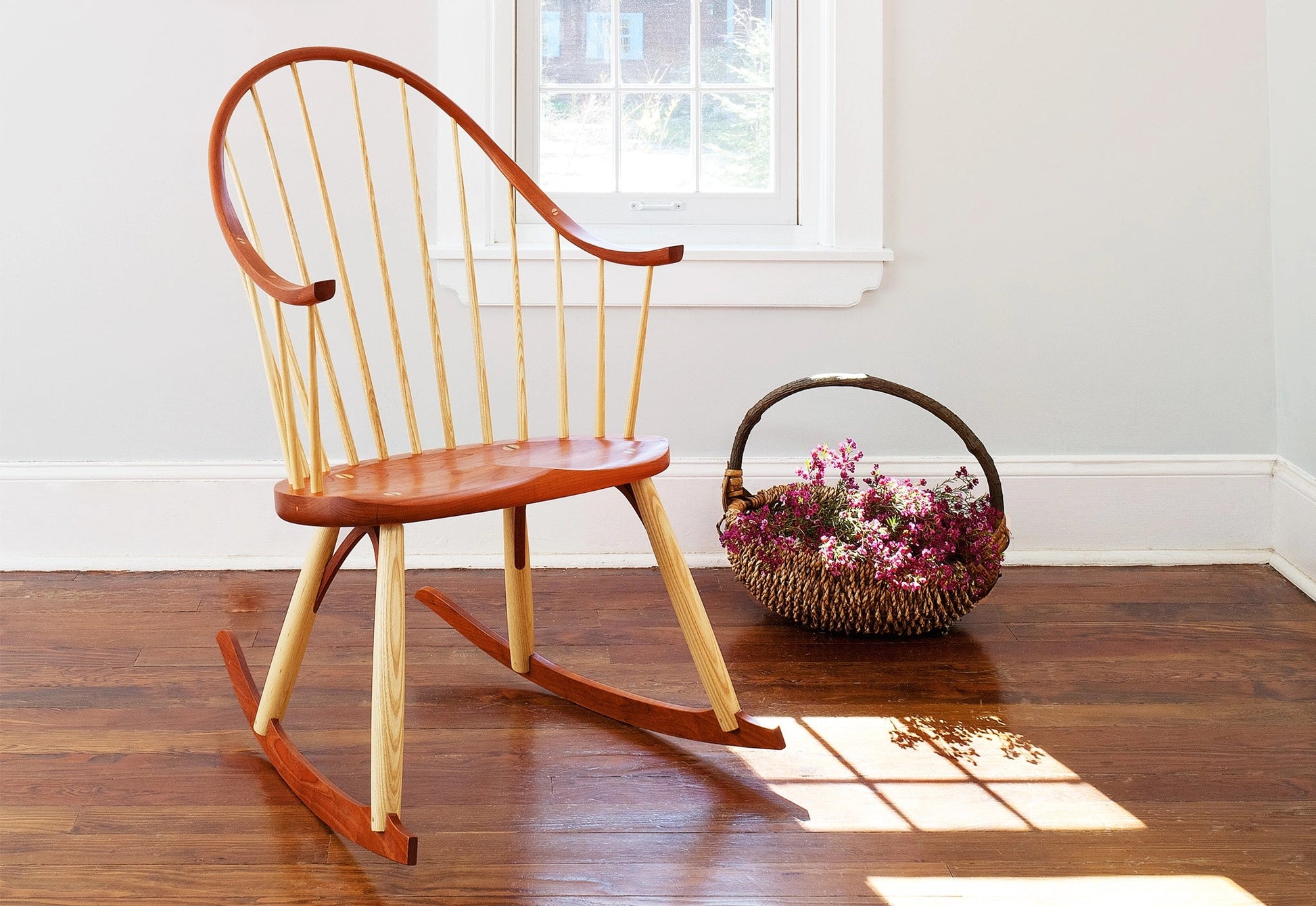 Wooden rocking chair on a wooden floor with a basket of flowers nearby.