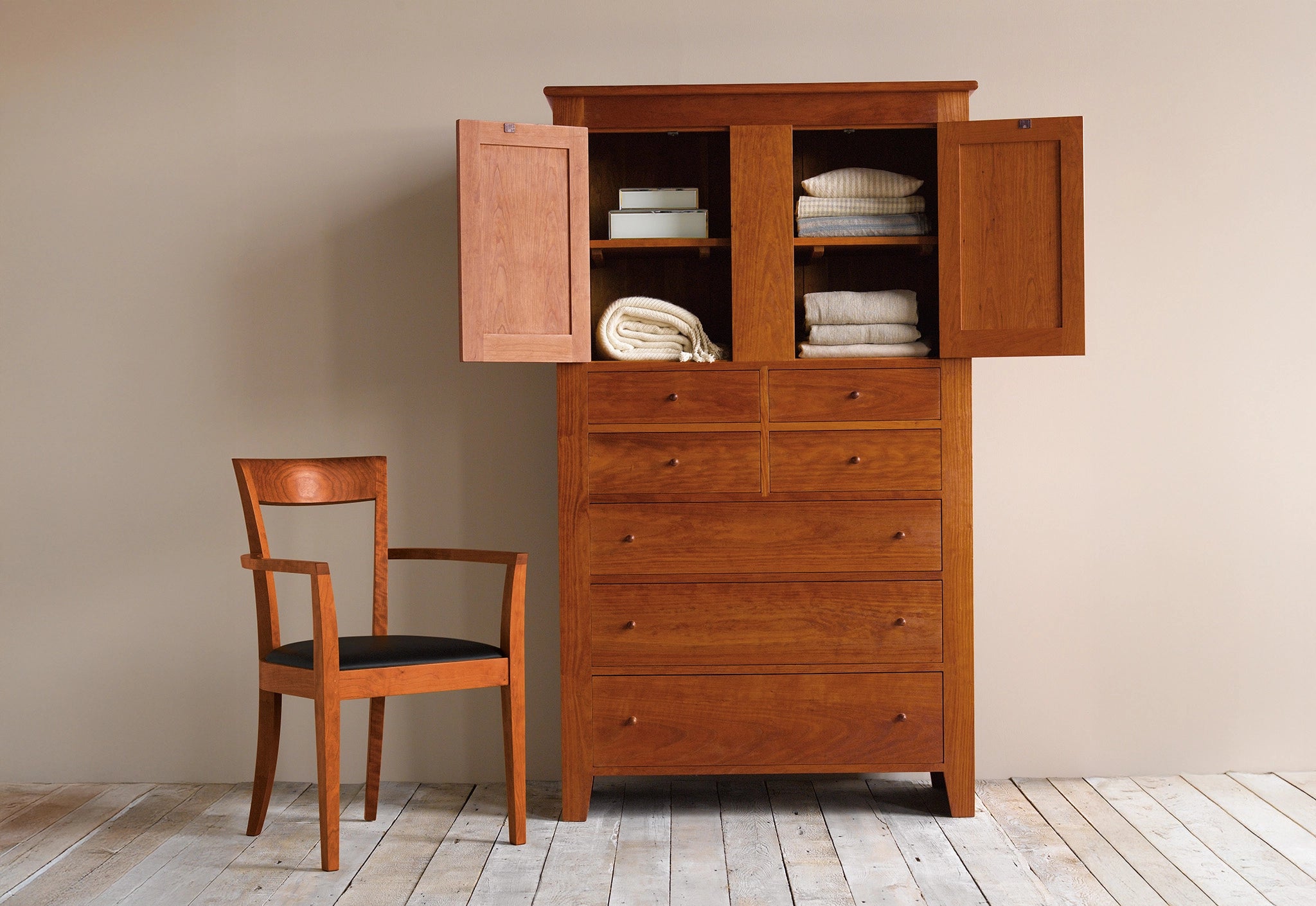 Wooden wardrobe with open doors revealing folded clothes, accompanied by a chair on a wooden floor.