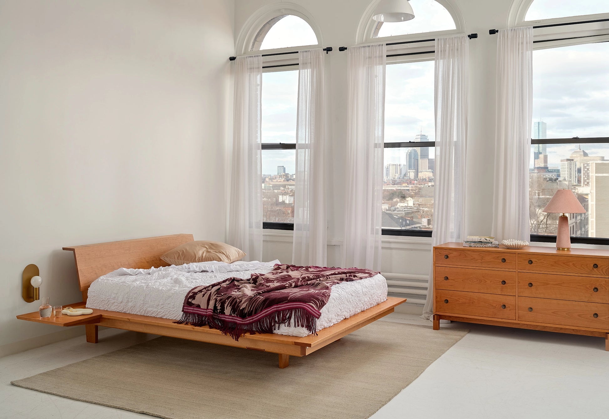 Modern bedroom with wooden platform bed and dresser, large windows with city view.
