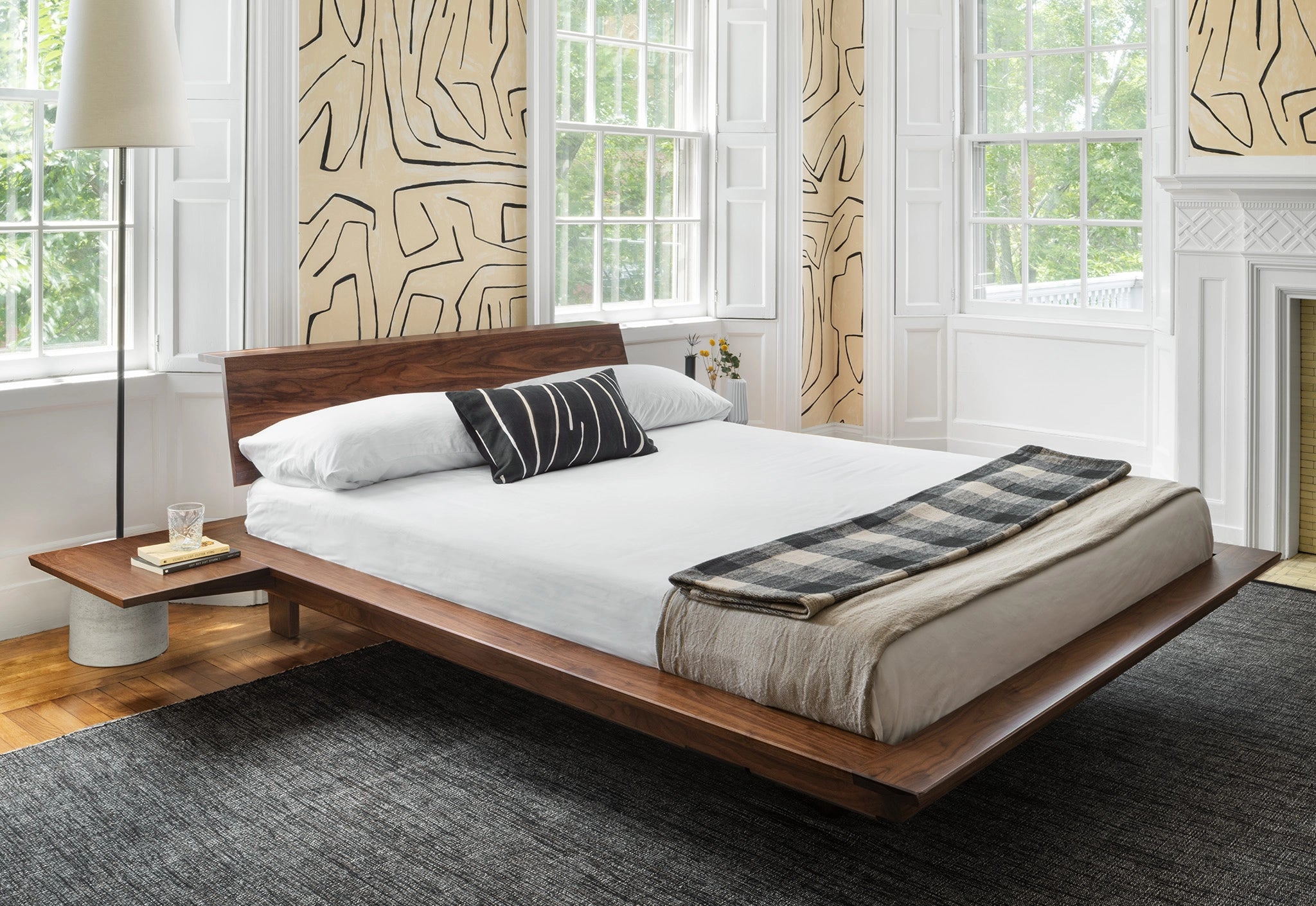 Wooden bed with white bedding in a room with large windows and patterned wallpaper.