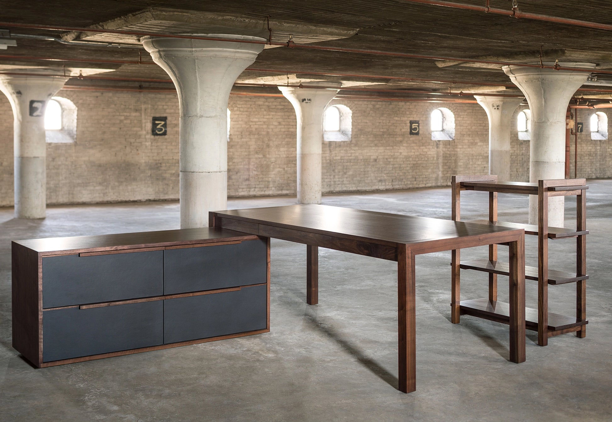 Modern office furniture including a desk and storage unit in an industrial-style room with concrete floors and exposed beams.