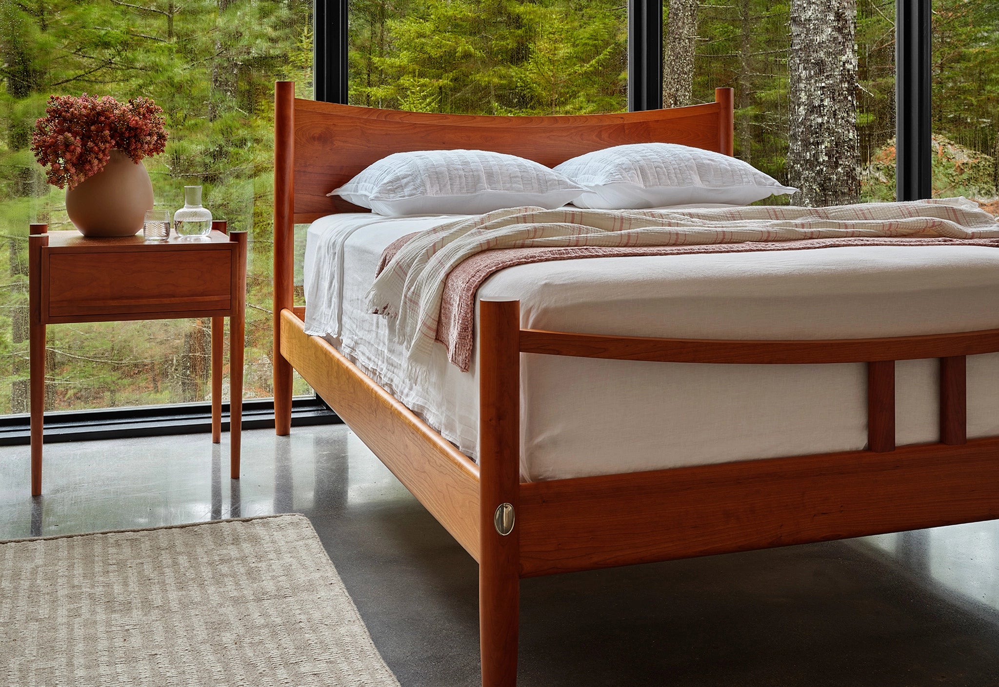 Wooden bed with white bedding in a room with large windows showing greenery