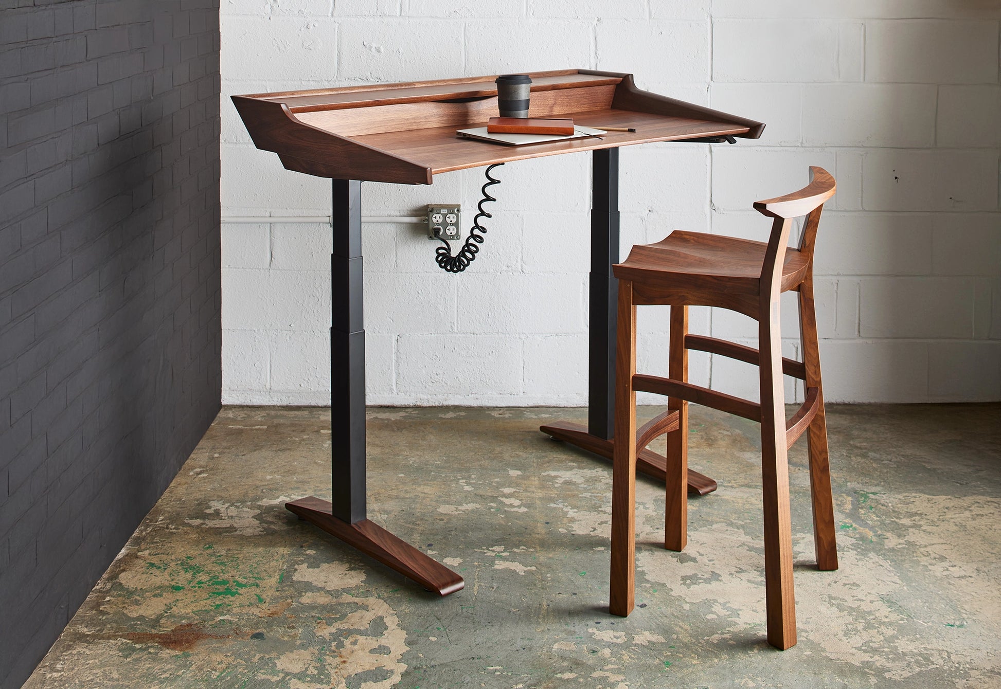 Wooden standing desk with a chair against a white brick wall.