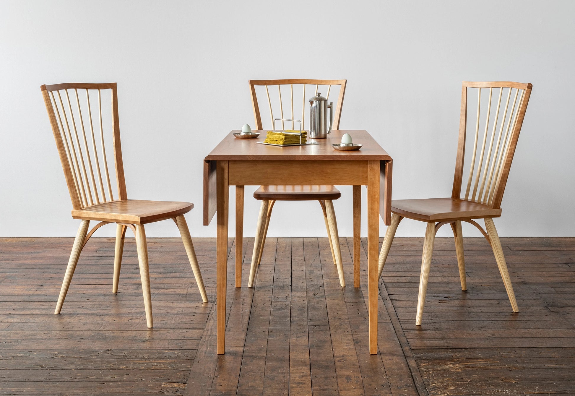 Wooden dining table with four chairs on a wooden floor against a white wall.