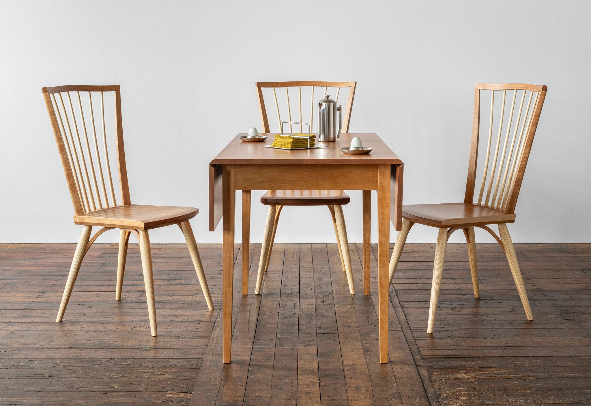 Wooden dining table with four chairs on a wooden floor against a white wall.