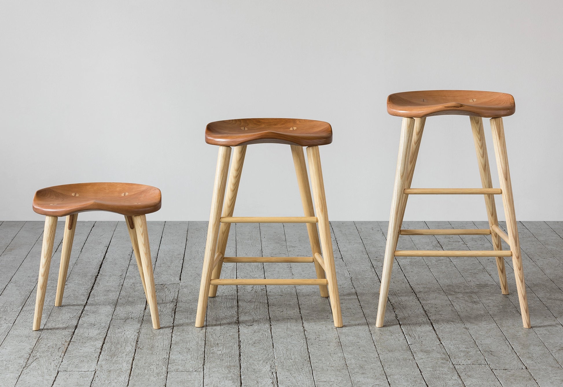 Three wooden stools on a gray wooden floor with a plain white wall background