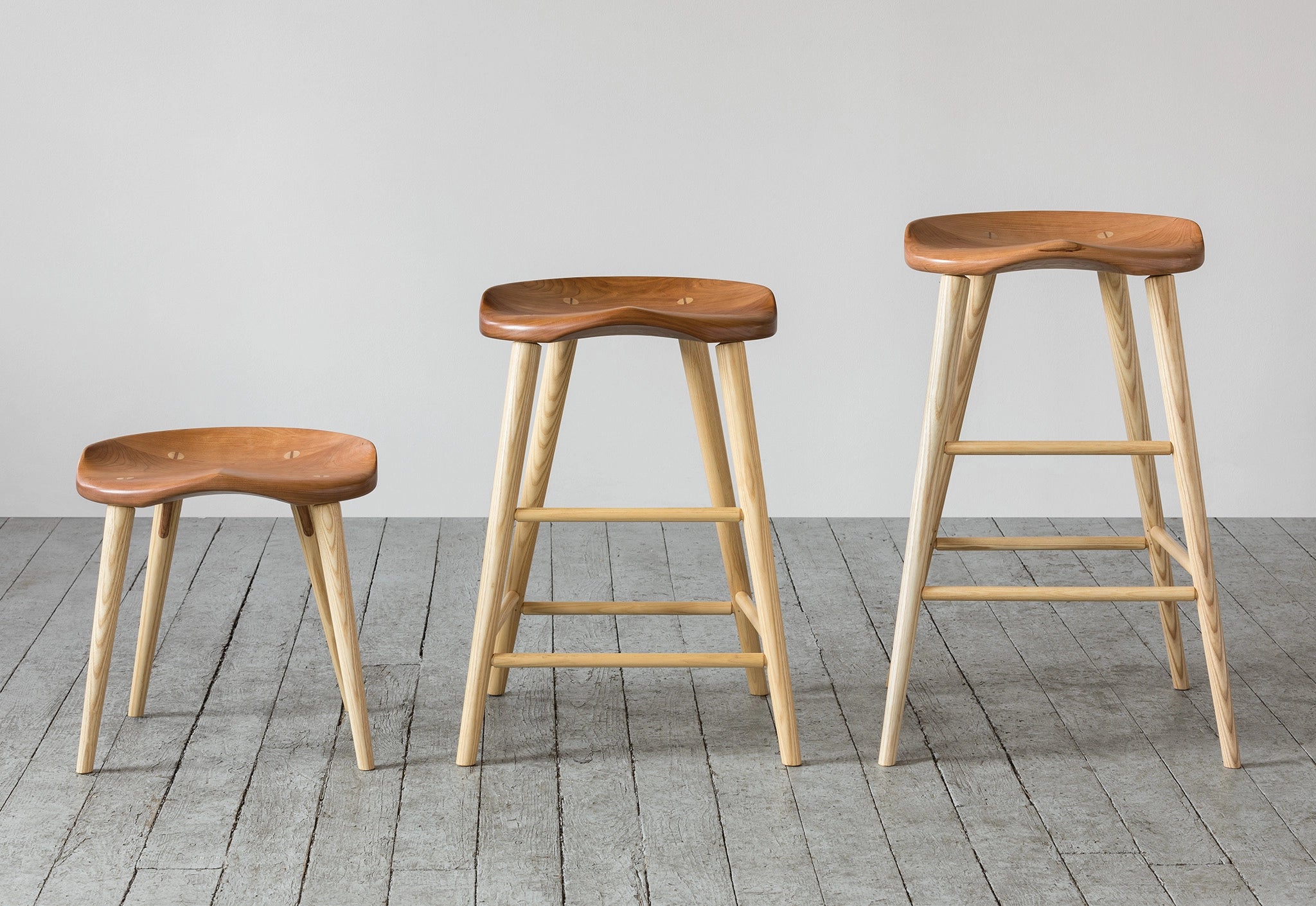 Three wooden stools on a gray wooden floor with a plain white wall background
