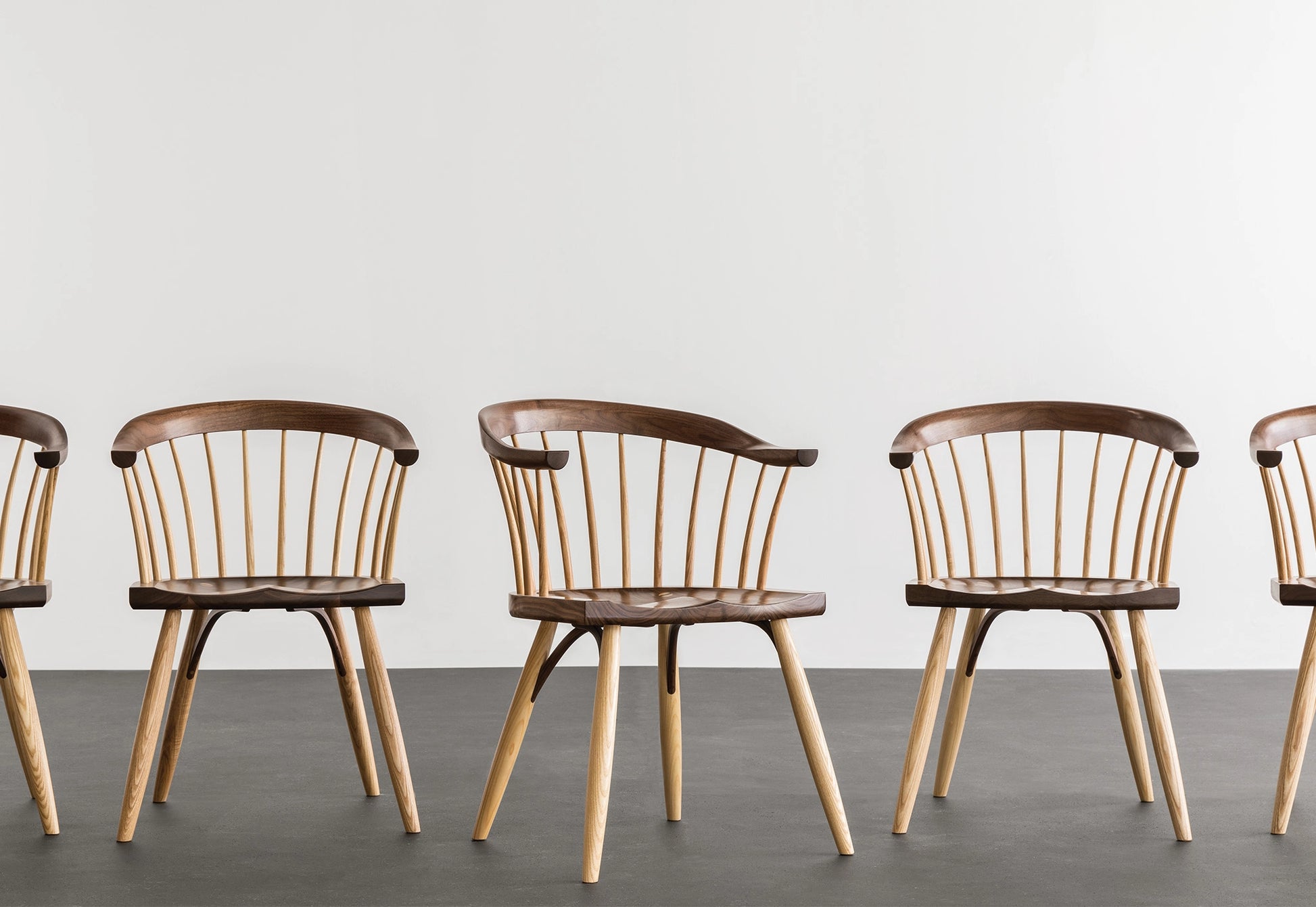 Row of wooden chairs with brown seats and light wood legs on a gray floor against a white wall.