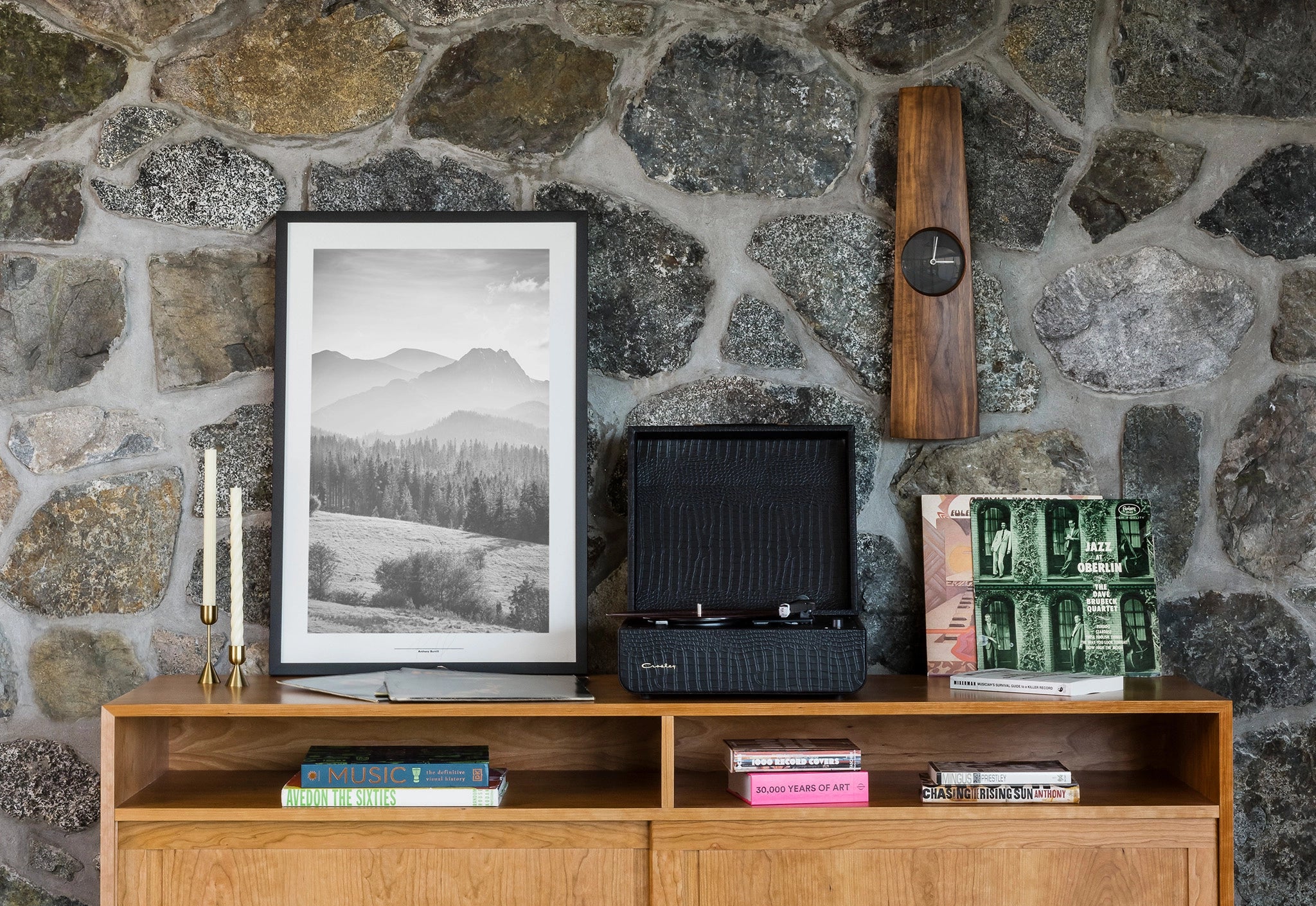 Wooden shelf against a stone wall with decorative items including a framed picture, clock, and books.
