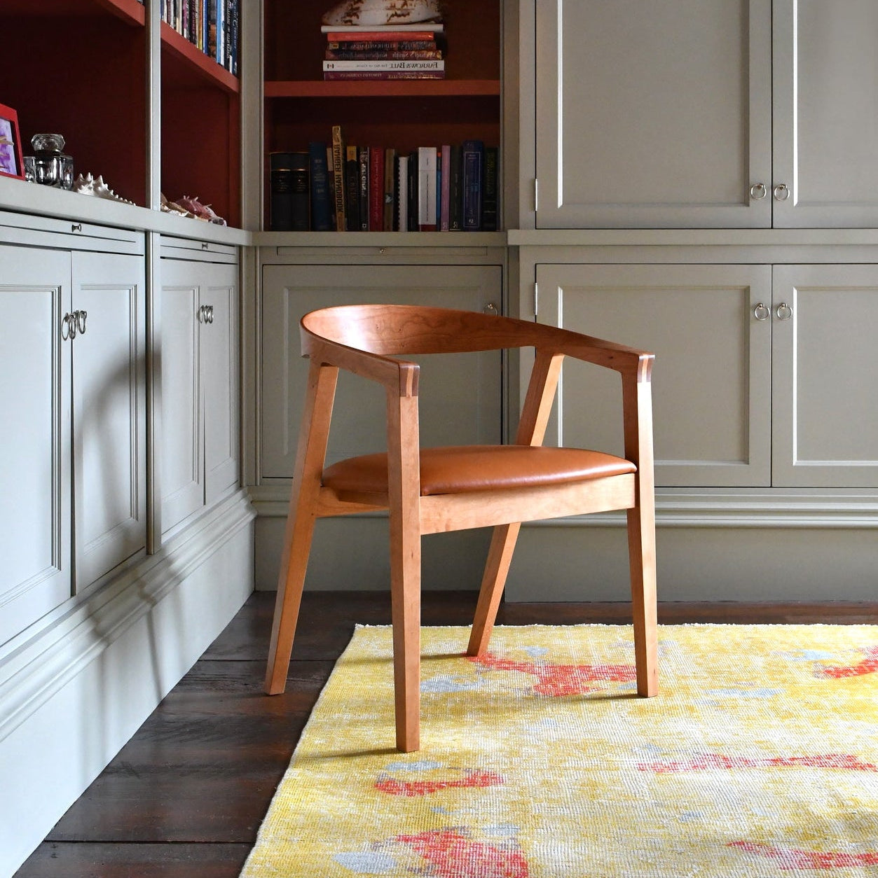 A mid-century modern wooden armchair with a curved back and light brown leather seat, positioned on a yellow patterned rug in front of built-in bookshelves and gray cabinets.