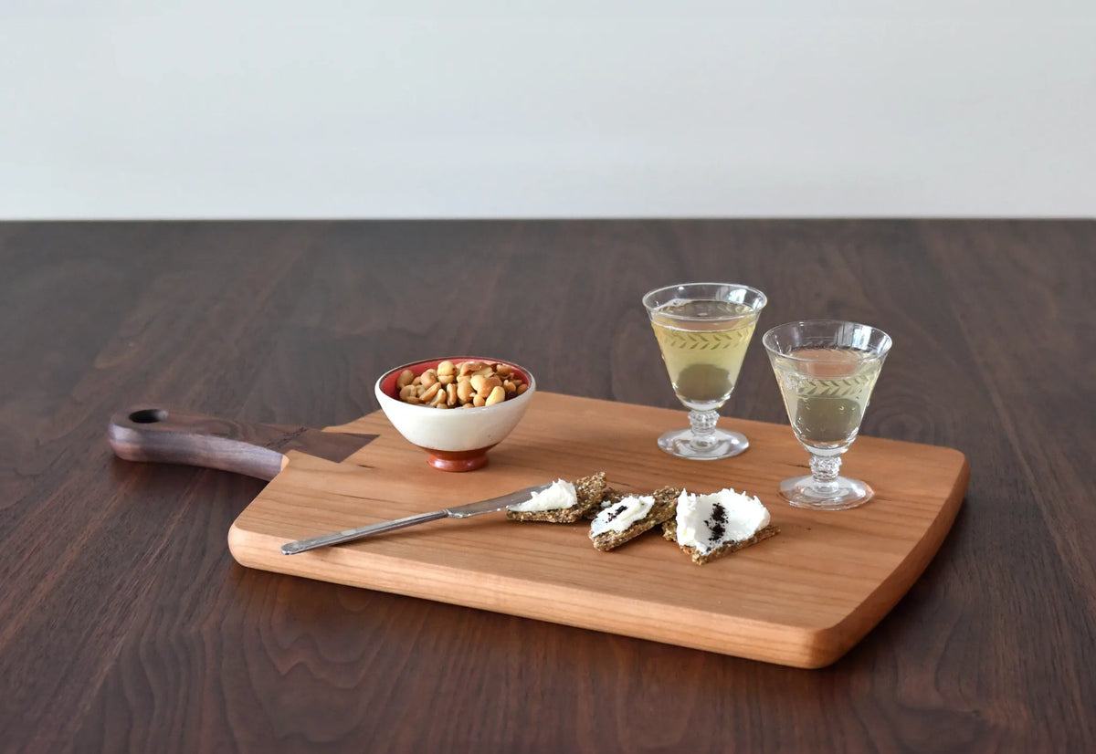 A rectangular wooden serving board with rounded corners and a dark handle, styled on a dark wood table with a bowl of nuts, cheese slices, and two small glasses of white wine.