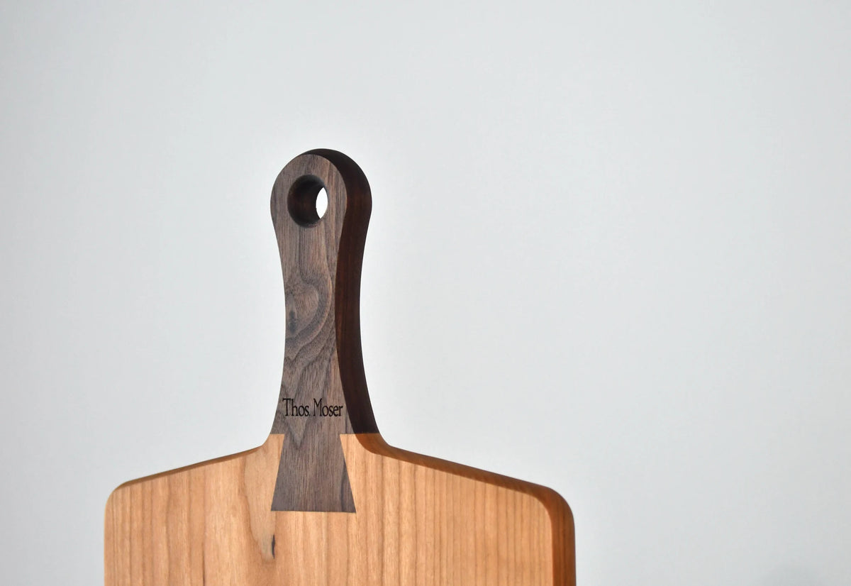 A wooden serving board with a dark wood handle featuring the engraved brand name 'Thos. Moore' and a circular hanging hole, set against a plain light gray background.