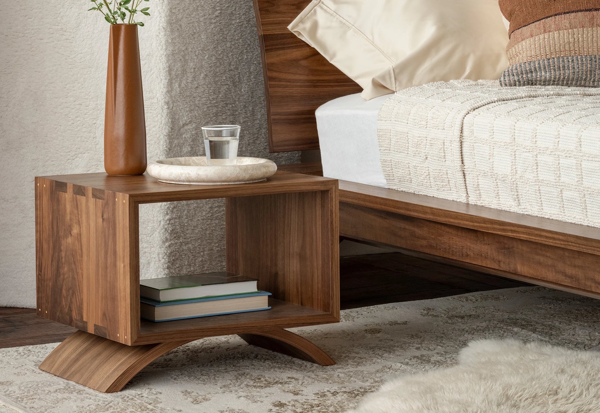 Wooden nightstand with books and a glass next to a bed with pillows and bedding.