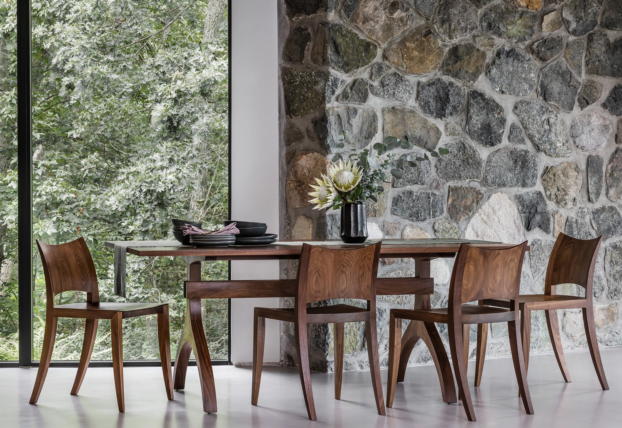 Dining area with wooden table and chairs against a stone wall with large windows showing greenery.