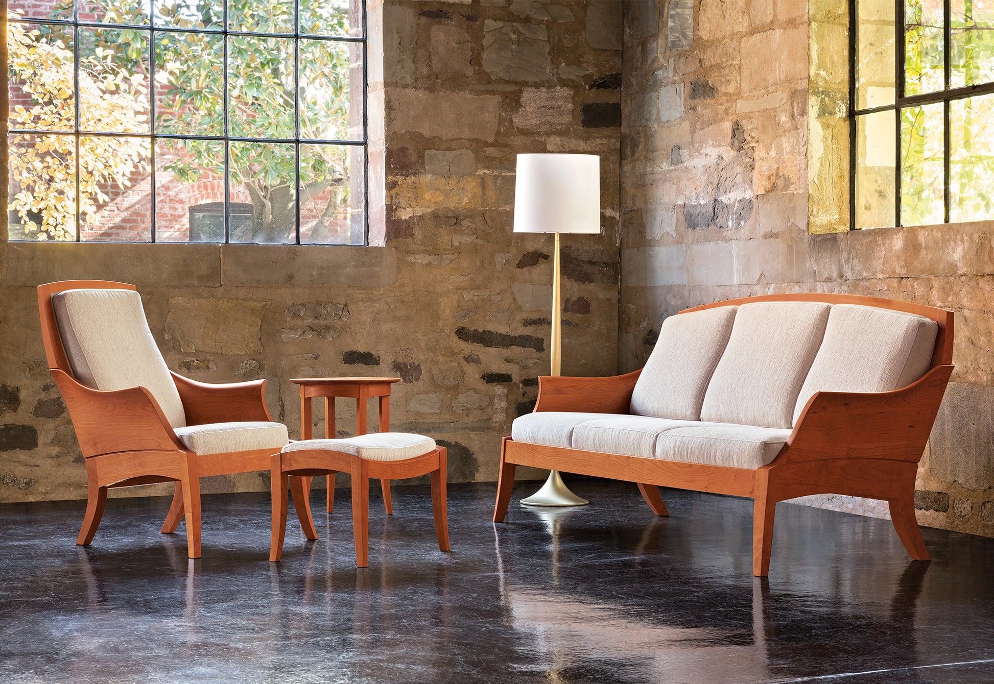 Wooden furniture set with a chair, sofa, and ottoman in a room with stone walls and large windows.