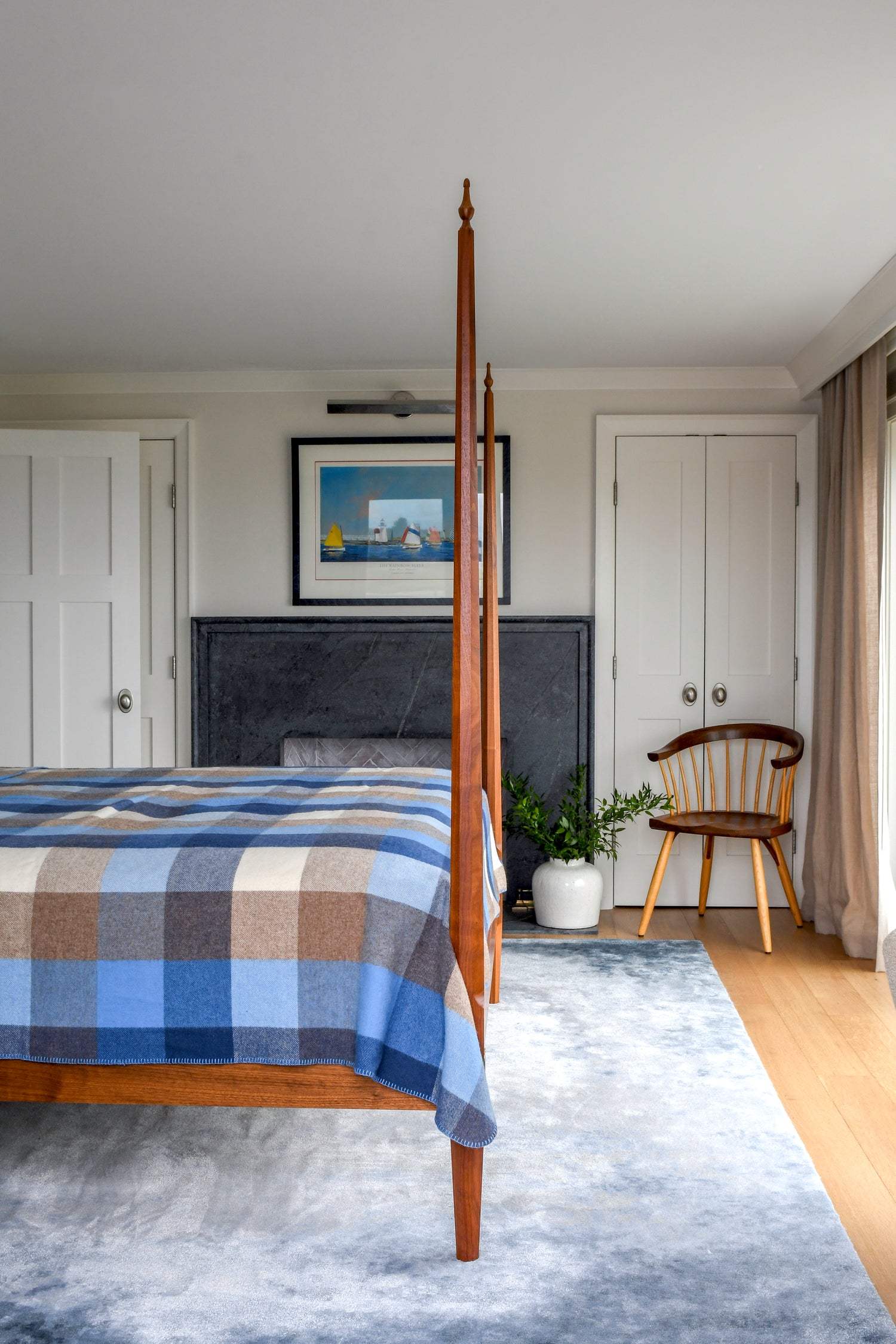 A bedroom scene featuring a wooden four-poster bed with a blue, brown, and beige plaid blanket, set in a modern room with a blue-gray rug, a wooden chair, and a dark fireplace.