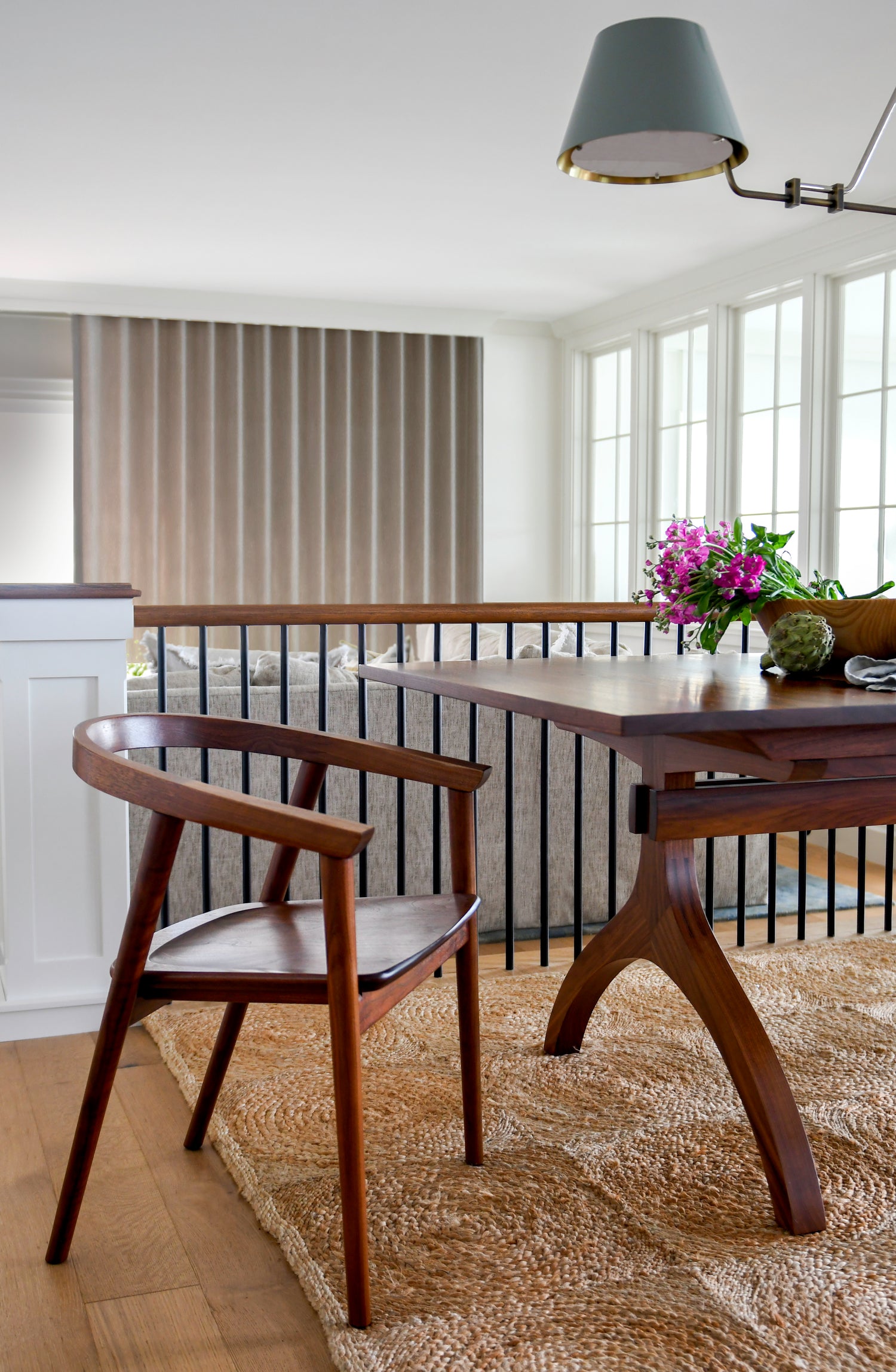 A mid-century modern wooden dining table and matching armchair in a bright, contemporary dining room with a jute rug, black metal railing, and large windows.
