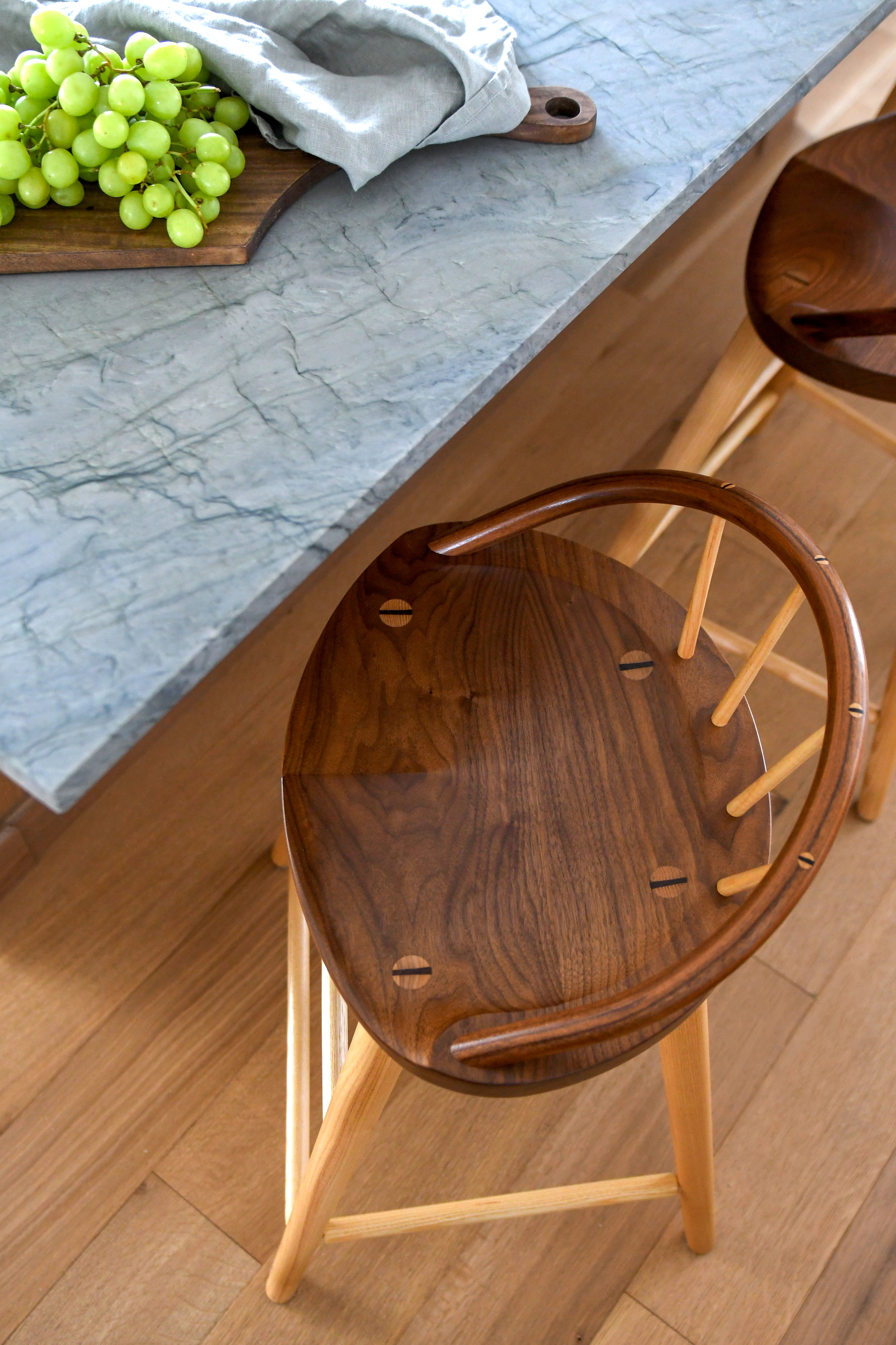 A close-up of a dark brown wooden bar stool with a curved backrest, positioned next to a gray stone countertop with green grapes and another stool partially visible.