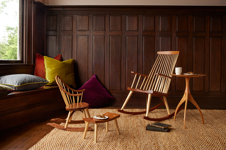 Children's sized rocking chair on left, small wooden footstool with a plate of cookies, New Glouceter Rocking chair on left beside a three-legged wooden side table in cherry with cup and flannel blankets and velvet pillows in background