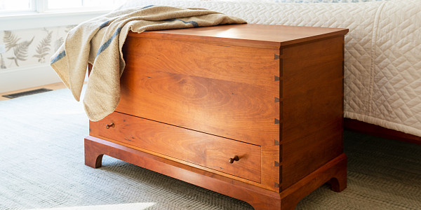 Blanket box in cherry sits at the foot of a bed wtih wool blanket draped over the edge