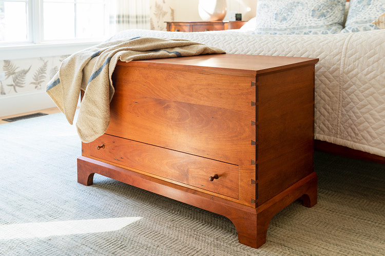 Blanket box in cherry sits at the foot of a bed wtih wool blanket draped over the edge