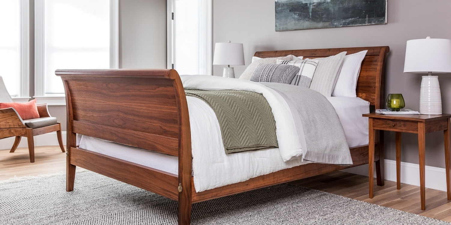 Sleigh Bed with curved footboard in walnut with square side table to the right and corner edge of wing lounge chair on left side