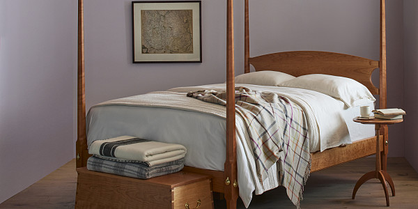 Bedroom with Pencil Post Bed in cherry with white linens and woolen blankets, Shaker round stand in cherry with books and coffee cup to right of bed, Captains Chest in cherry with blankets folded on top sits at the foot of the bed. Picture of a map hangs on the wall