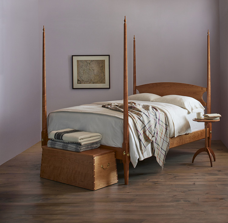 Bedroom with Pencil Post Bed in cherry with white linens and woolen blankets, Shaker round stand in cherry with books and coffee cup to right of bed, Captains Chest in cherry with blankets folded on top sits at the foot of the bed. Picture of a map hangs on the wall