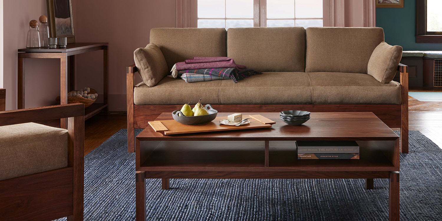 Living room with pink walls, walnut sofa with cushions and blankets, coffee table in walnut with cherry tray fruit and cheese, console table in back left with water vase and ceramic cups, and wooden lounge chair in front left with wooden arms and upholstery
