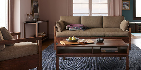 Living room with pink walls, walnut sofa with cushions and blankets, coffee table in walnut wiht cherry tray fruit and cheese, console table in back left with water vase and ceramic cups, and wooden lounge chair in front left with wooden arms and upholstery