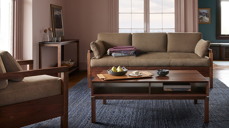 Living room with pink walls, walnut sofa with cushions and blankets, coffee table in walnut wiht cherry tray fruit and cheese, console table in back left with water vase and ceramic cups, and wooden lounge chair in front left with wooden arms and upholstery