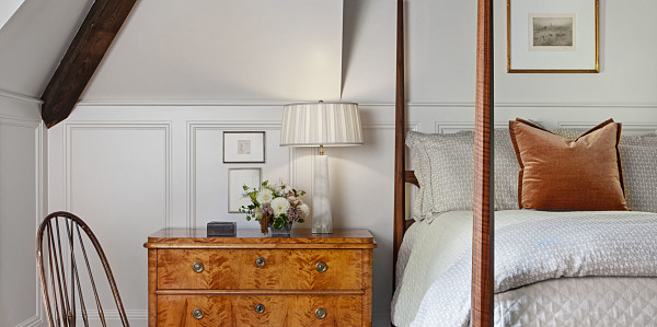 Bedroom with walnut pencil post bed, vintage burl dresser and windsor chair. Exposed timber beams with white rug.