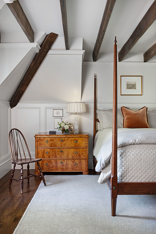Bedroom with walnut pencil post bed, vintage burl dresser and windsor chair. Exposed timber beams with white rug.