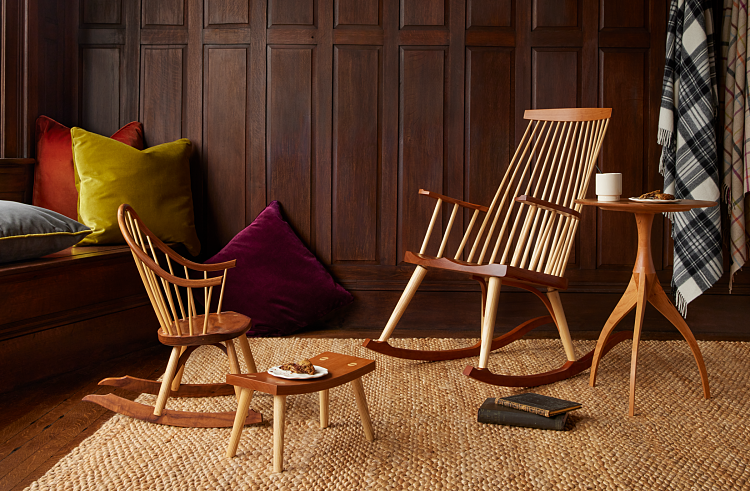 Children's sized rocking chair on left, small wooden footstool with a plate of cookies, New Glouceter Rocking chair on left beside a three-legged wooden side table in cherry with cup and flannel blankets and velvet pillows in background