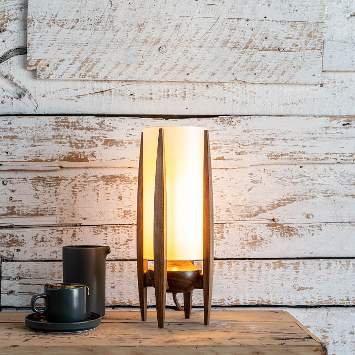 Mid-Century style lamp with a tea cup and pitcher beside it, sitting on a wooden table in front of a white wall.