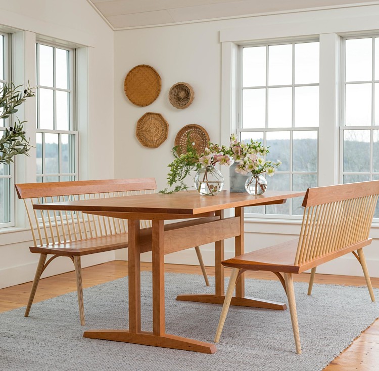 Eastward bench and trestle table in cherry featured in a dining room with vase of flowers and baskets on wall