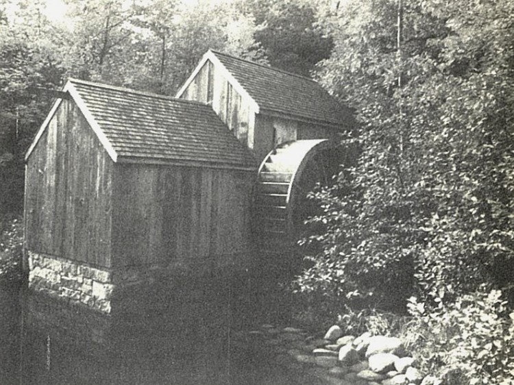 black and white image of an old water house and water wheel next to a pond set within trees