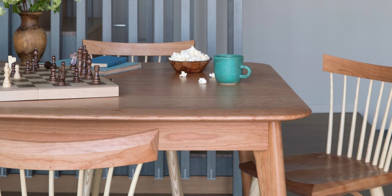 A Wooden table in cherry surrounded by spindle chairs in cherry and ash. On the table there is an aquamarine coffee mug, a bowl of popcorn, and a chess board.