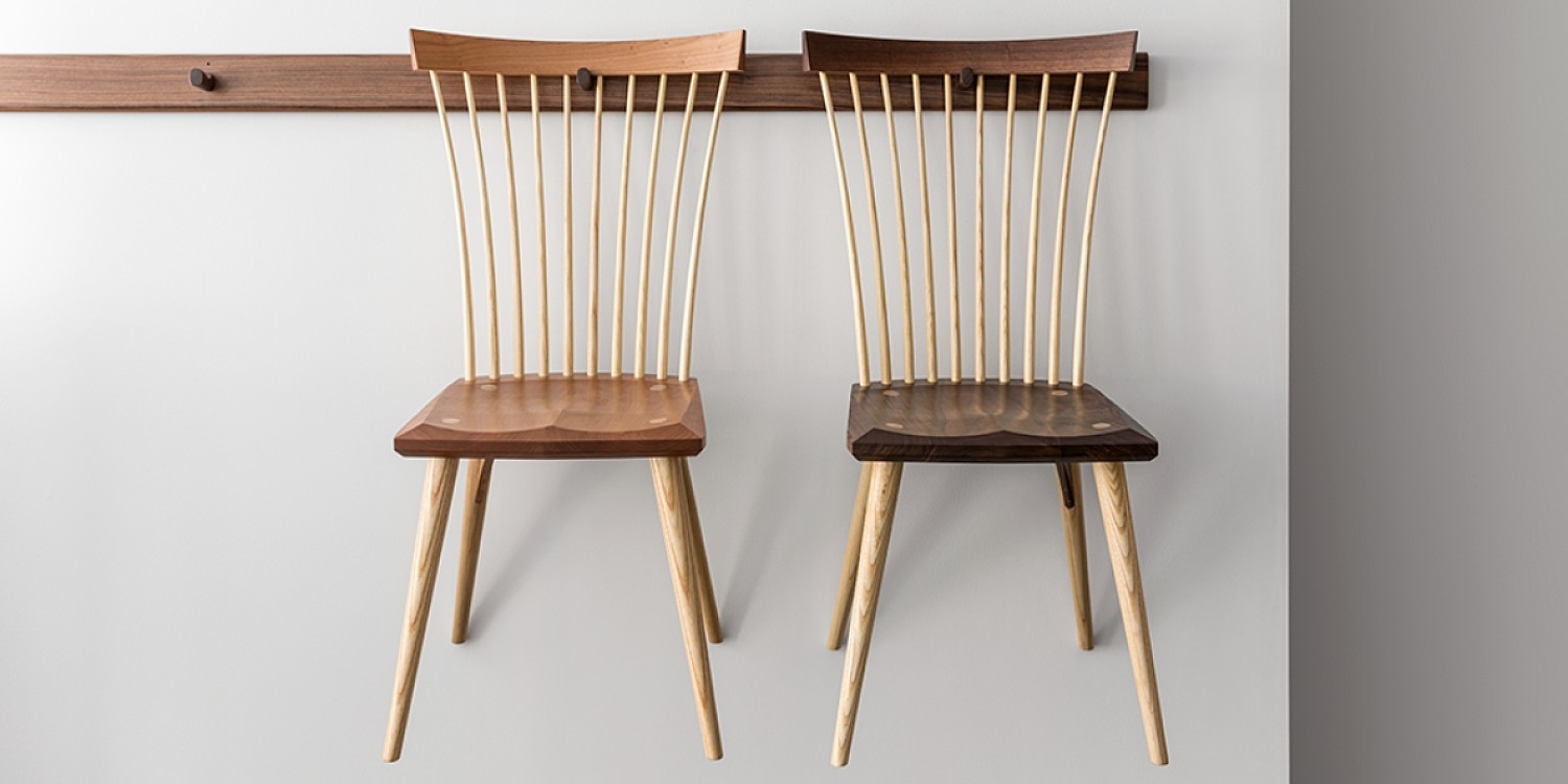 Two wooden chairs (Eastward Chairs) one in cherry one in walnut hang on a shaker wall post.