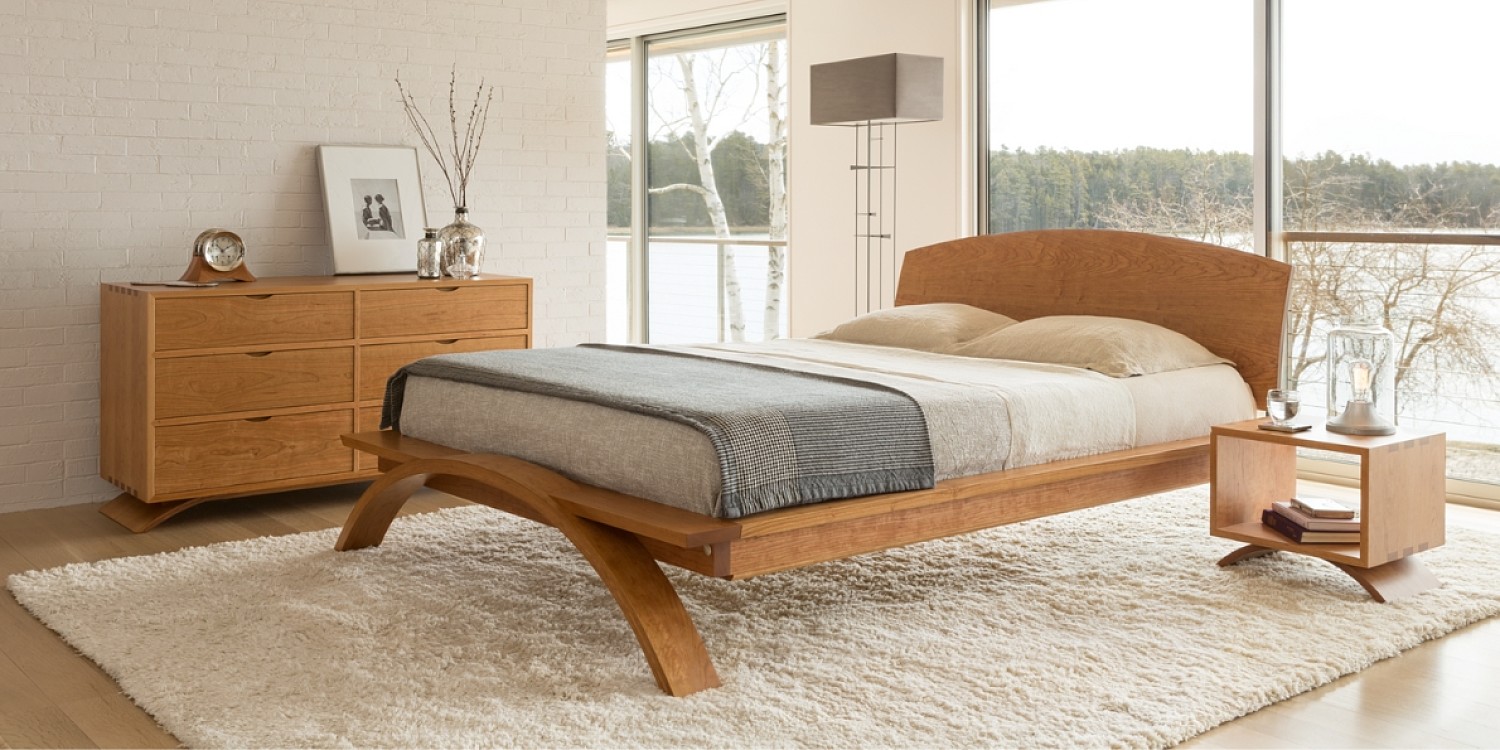 A styled bedroom with floor to ceiling windows to the right of the room. There is a solid cherry wood platform bed in the center featuring a curved foot and headboard with a grey blanket and white sheets and pillows. To the right of the bed is a small wooden vita cube side table with a lamp and glass of water on top and books on the lower shelf. To the left of the bed is a wooden dresser that has the same curved foot as the bed and features six drawers with recessed pulls. There is a clock, vases and black and white photograph perched on top of the dresser. the bed and side table are on a white shaggy rug.