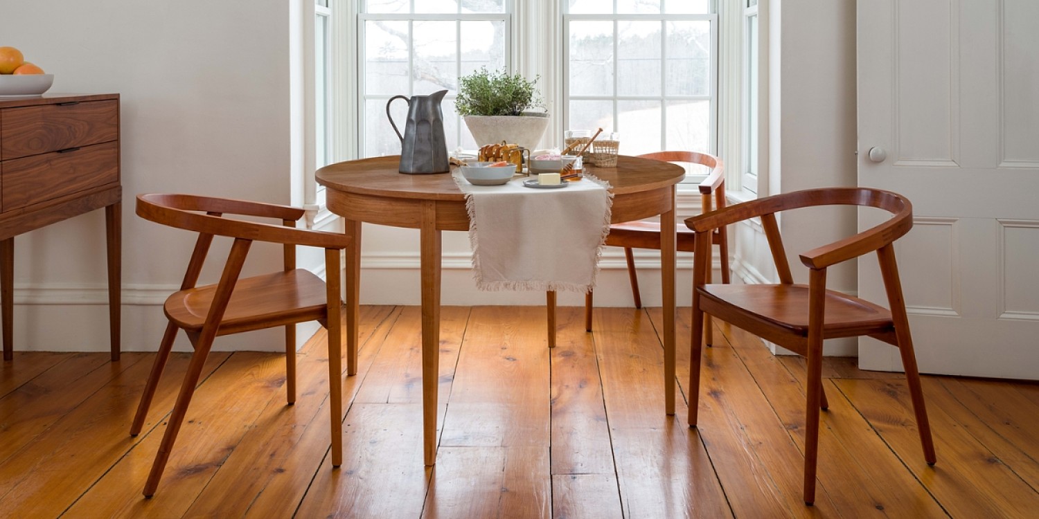 Breakfast nook with a round table and three wooden chairs with a rounded crest and arms in cherry at the table. A walnut sideboard is to the left with a bowl of oranges on top. The table is dressed with a white linen runner, black pitcher, herbs and dish ware.