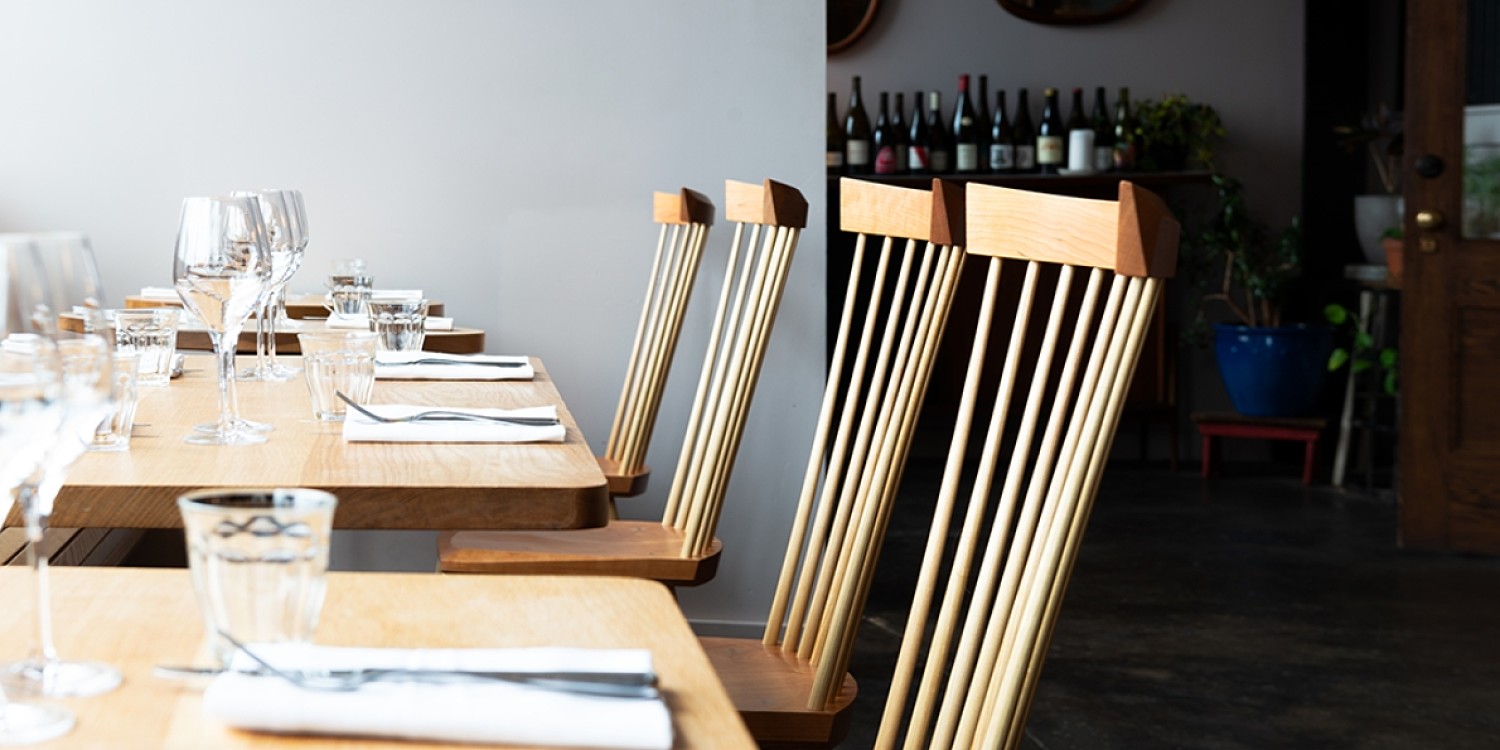 Four spindle chair crests are beside wooden tables in a restaurant. There are white cloth napkins, sliver ware and water glasses on the table.