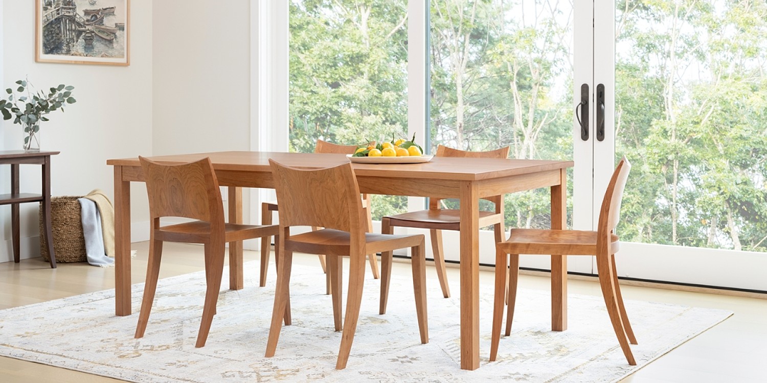A dining room wiht a white carpet and floor to ceiling windows that look out to a wooded lot. There is a square cherry dining table and four cherry dining chairs with solid backs around the table. On the table is a shallow dish with lemons and napkins. On the left wall there is a walnut hall table and a basket with blankets on the ground. There is a nautical piece of art that hangs on the wall above the hall table.